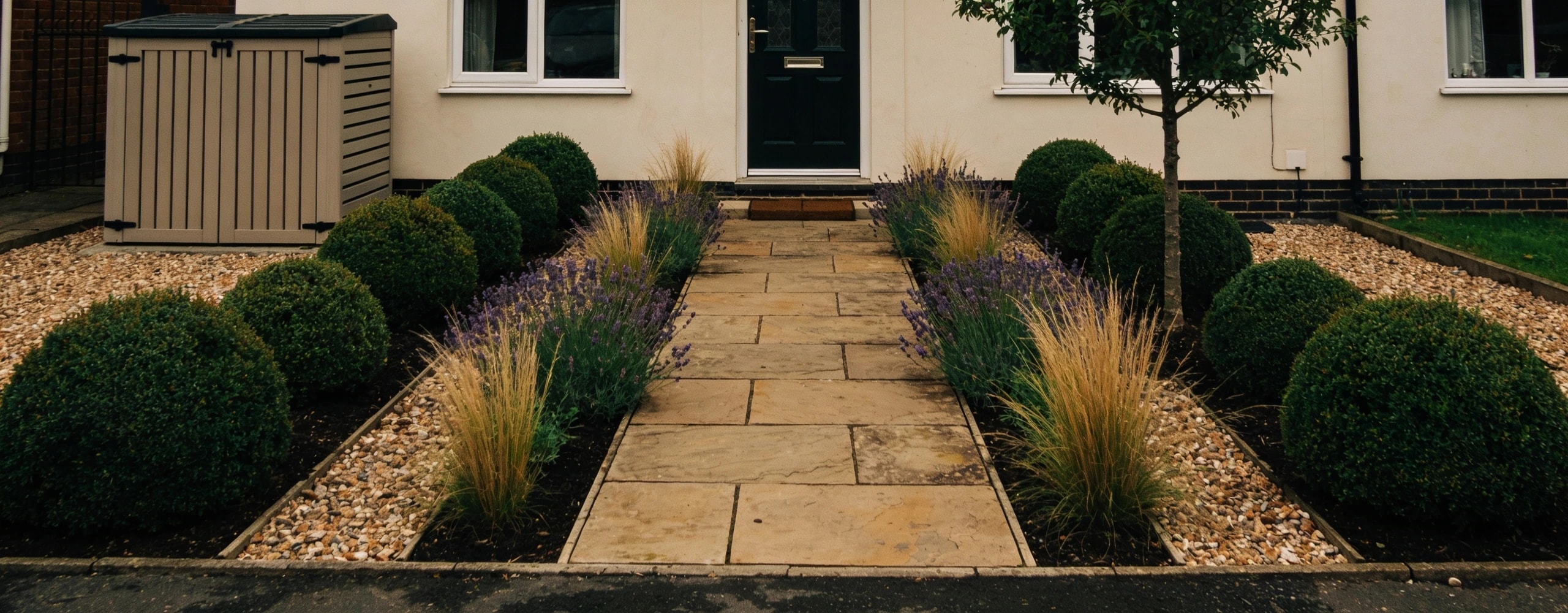 A front garden built around one route and repeated planting Small front garden with one wide stone path, deep beds of clipped evergreens, lavender and grasses