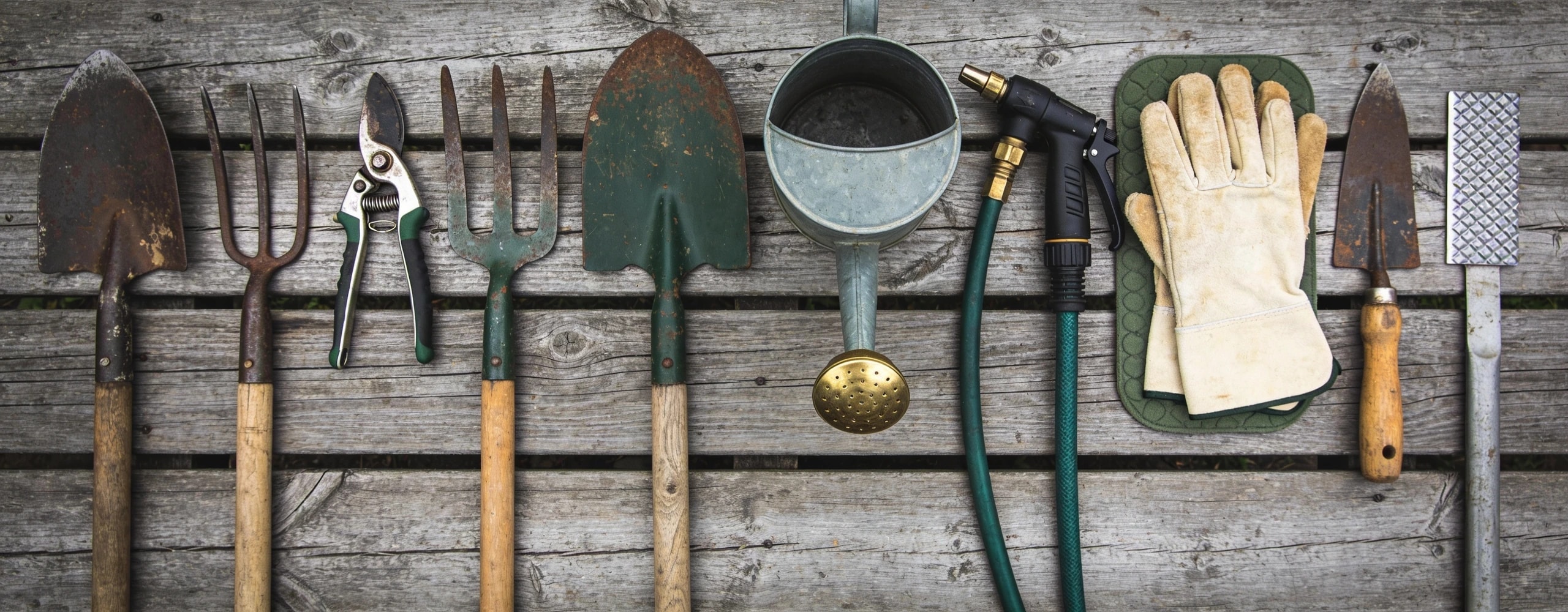 Twelve used garden hand and long tools laid out in a row on weathered timber boards